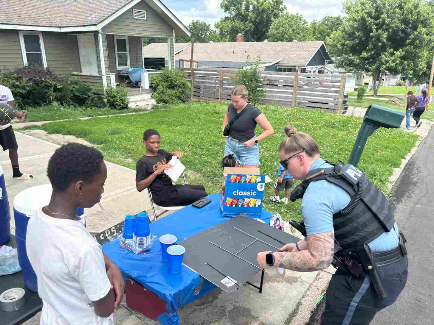 A family stands on a front lawn, as a police officer fixes a sign for a lemonade stand run by two young Black boys