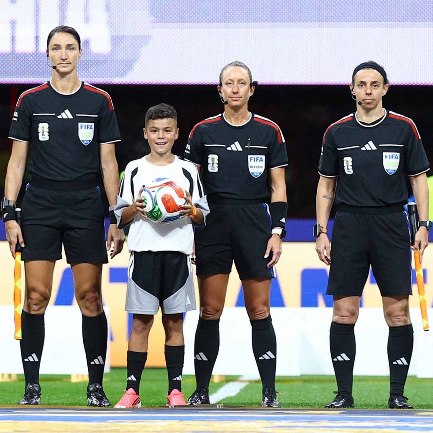 Three female match officials in black FIFA referee kits line up on the pitch beside a young mascot holding the match ball.