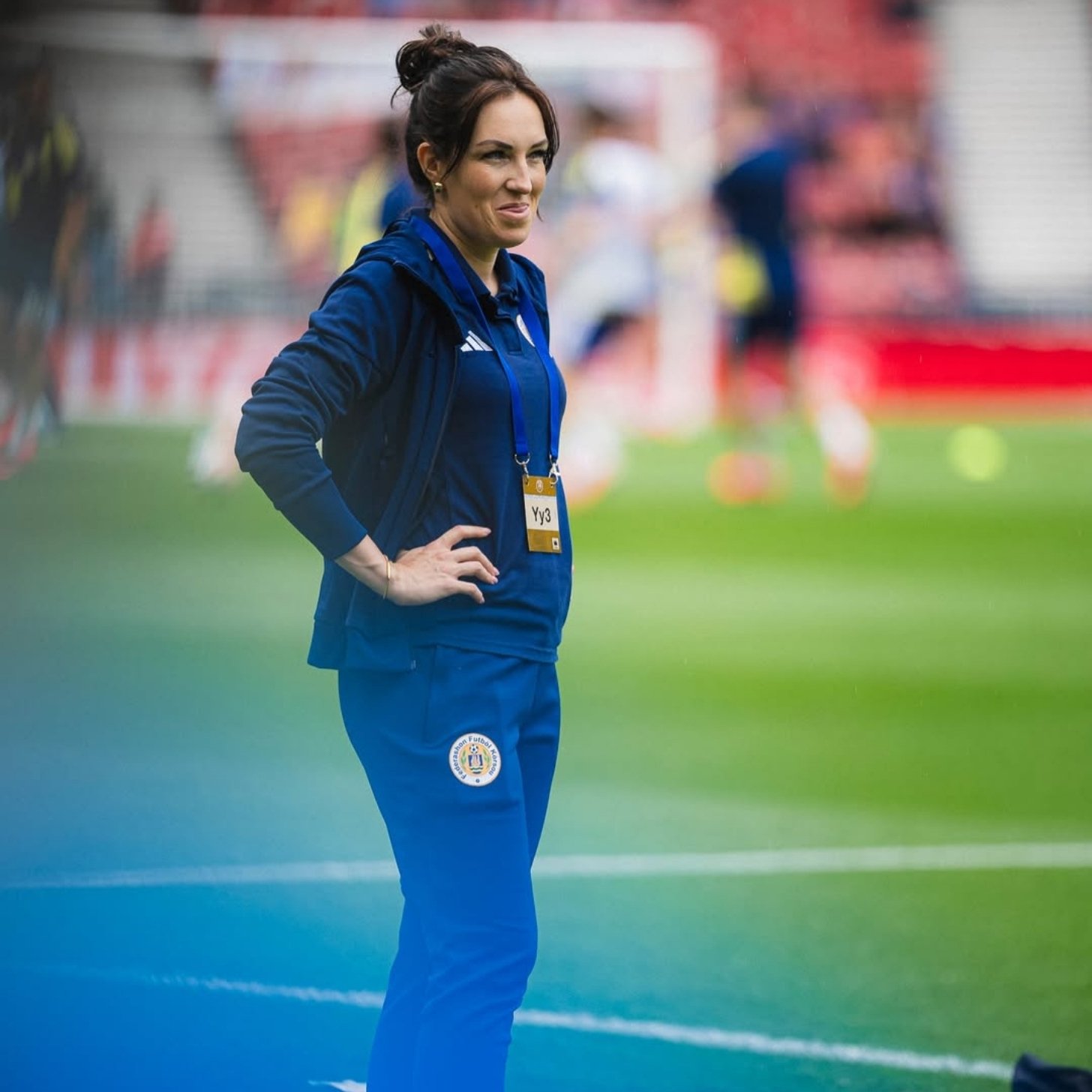 A woman in a navy Adidas tracksuit stands with her hands on her hips beside a football pitch, wearing an accreditation lanyard.