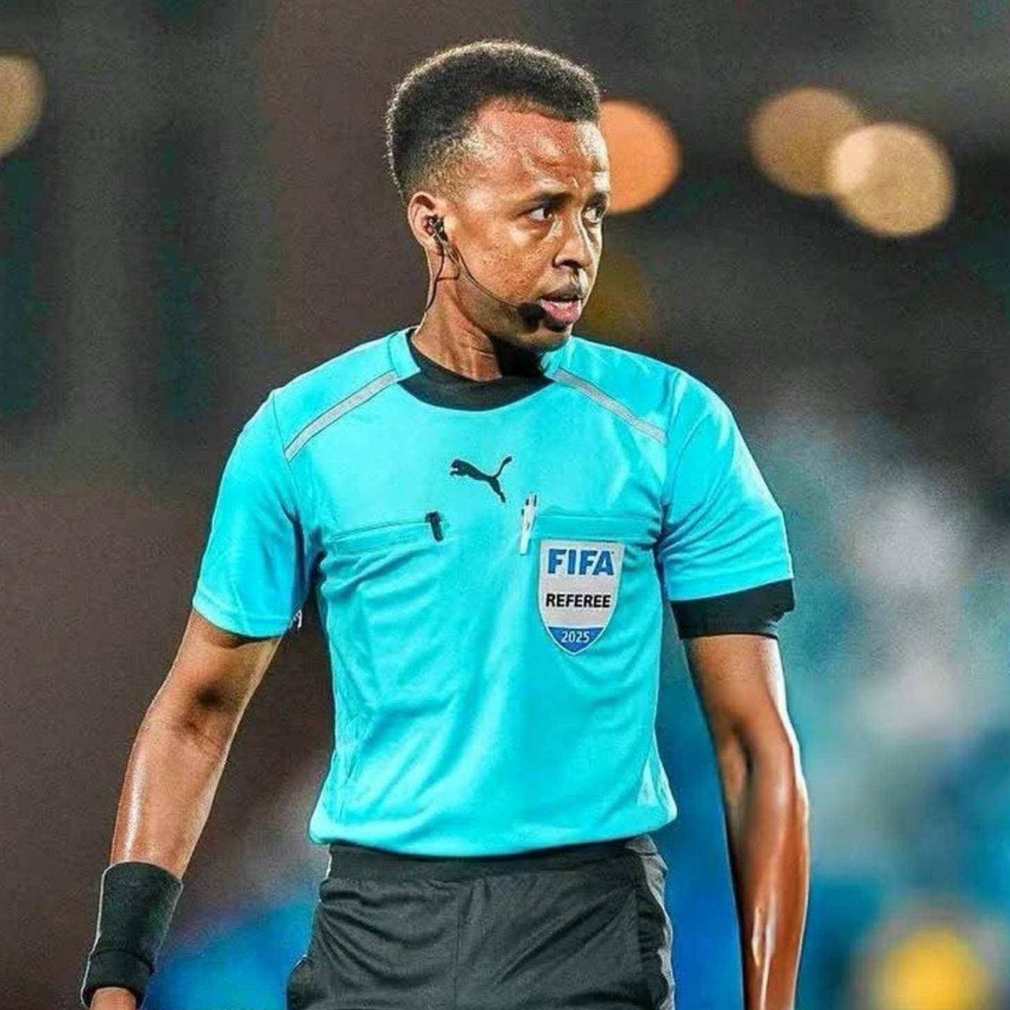 A football referee in a teal FIFA referee jersey walks across a floodlit pitch wearing an earpiece and microphone.