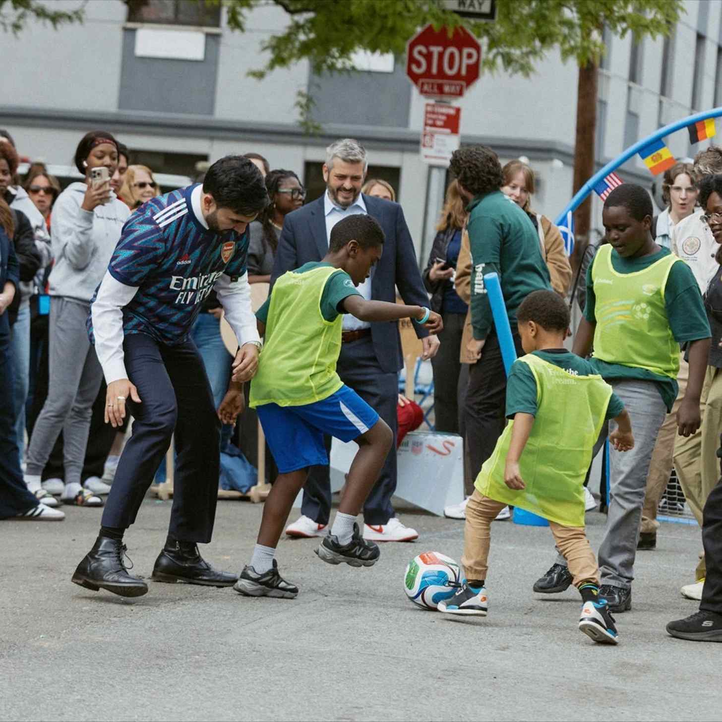 A man in an Arsenal shirt plays street football with children in yellow bibs while a crowd watches and films, with national flags strung overhead.