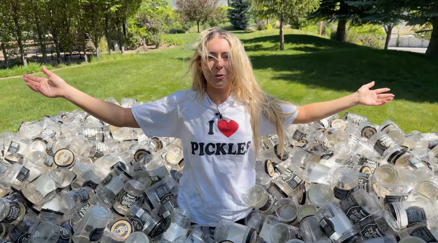 A woman stands in a massive pile of pickle jars, wearing a shirt that reads I Love Pickles
