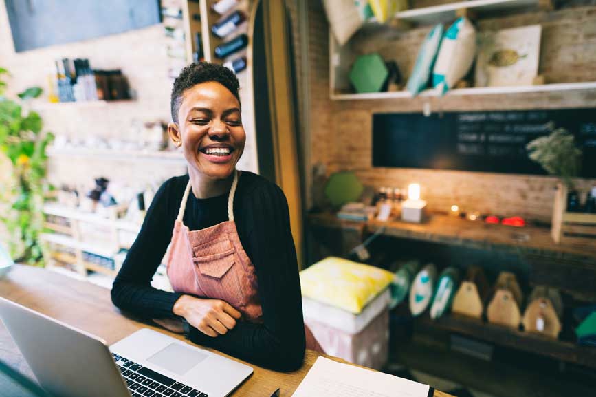 A Black woman with short hair smiles behind her computer in her small business / retail shop
