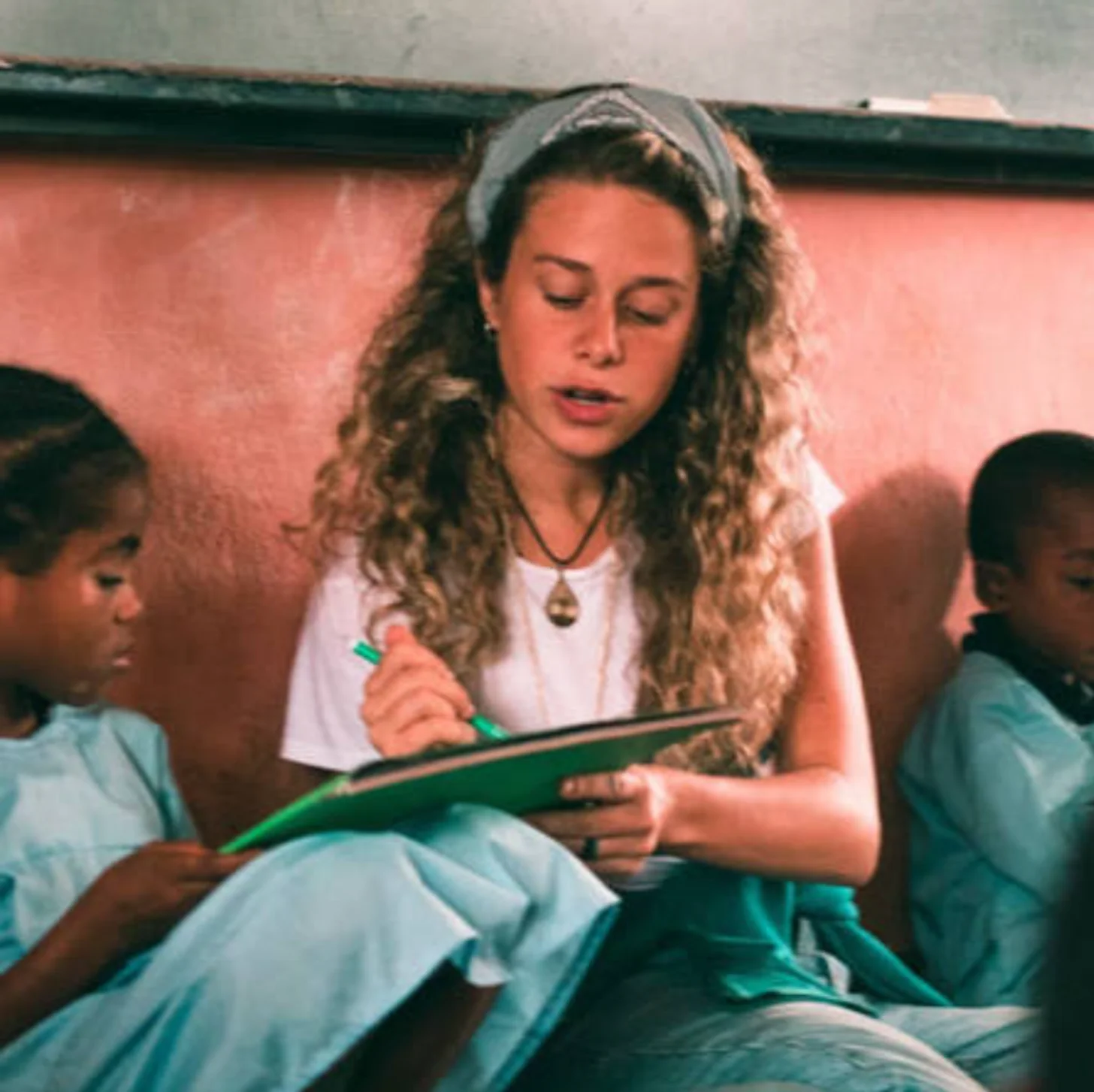 A woman with curly hair and a headband reads from a green clipboard to a group of children seated beside her against a coral-colored wall.