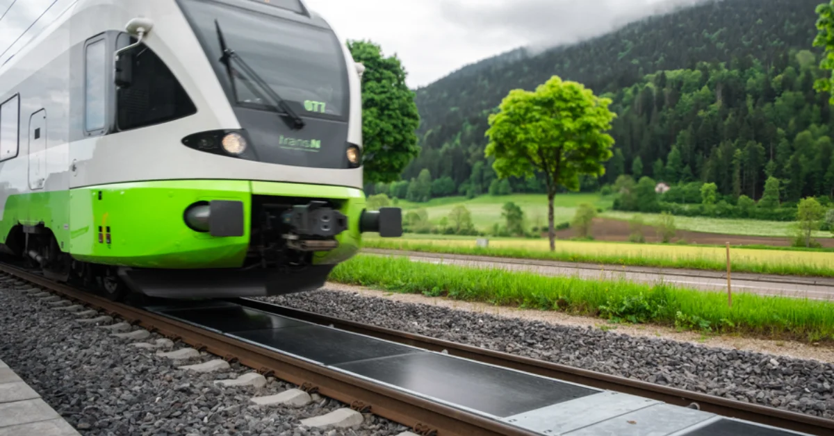 A modern green and white electric train travels through a lush valley with forested mountains, with solar panels on the train tracks