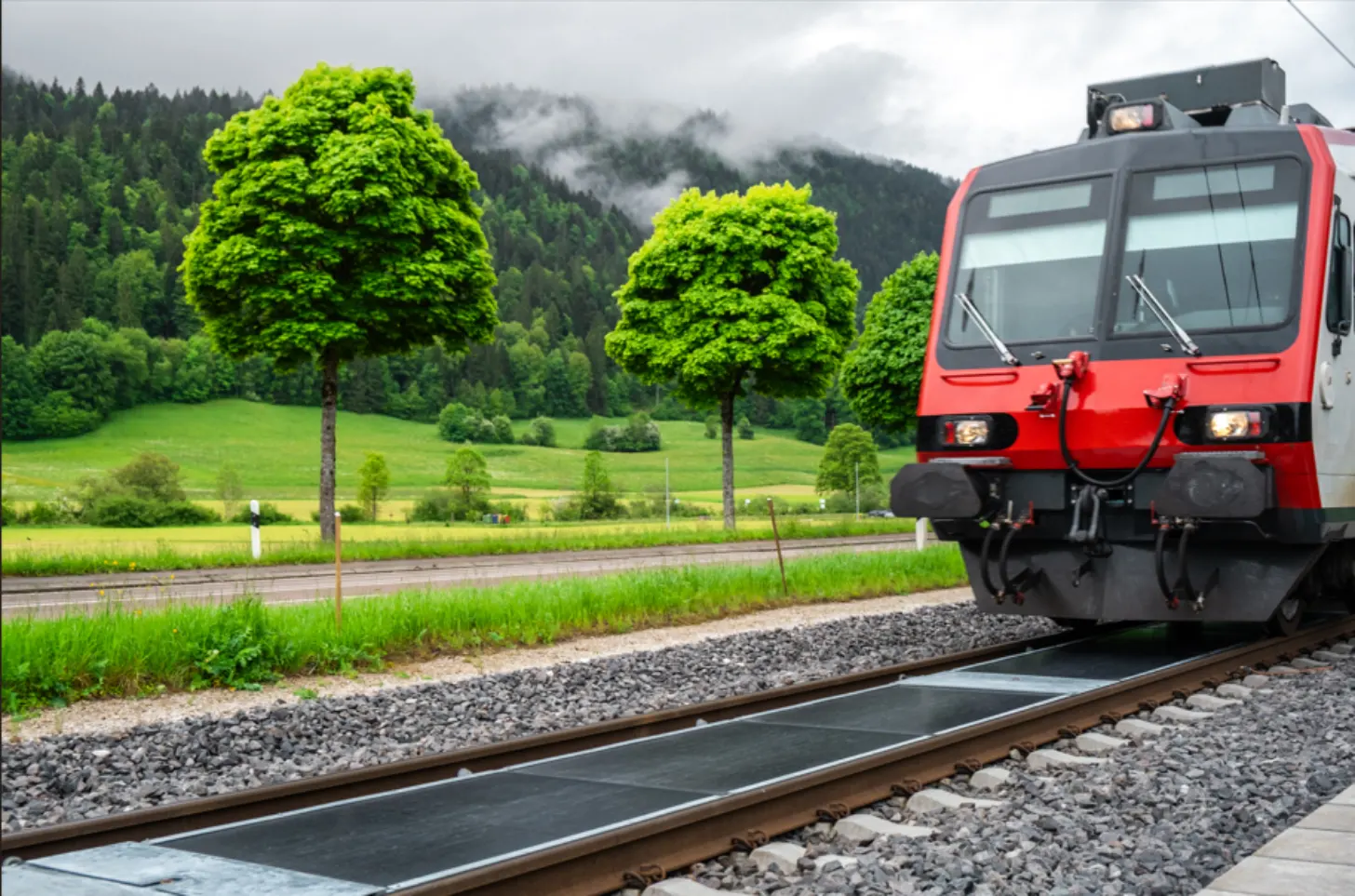 A red commuter train travels through a lush alpine valley over train tracks with solar panels between the rails