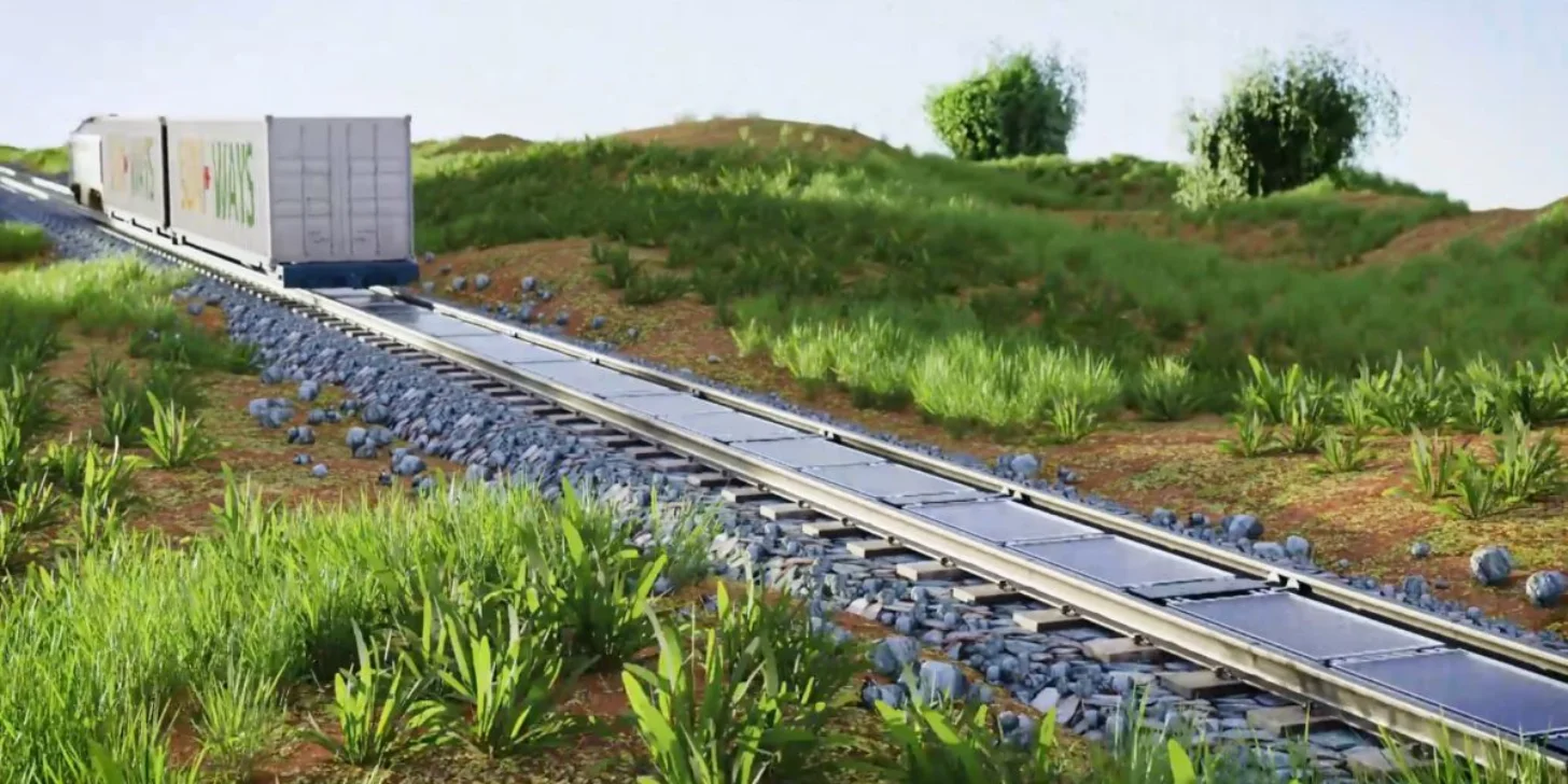 A freight train carrying cargo containers travels along an elevated rail line through a lush, green hillside landscape,with solar panels on the train tracks