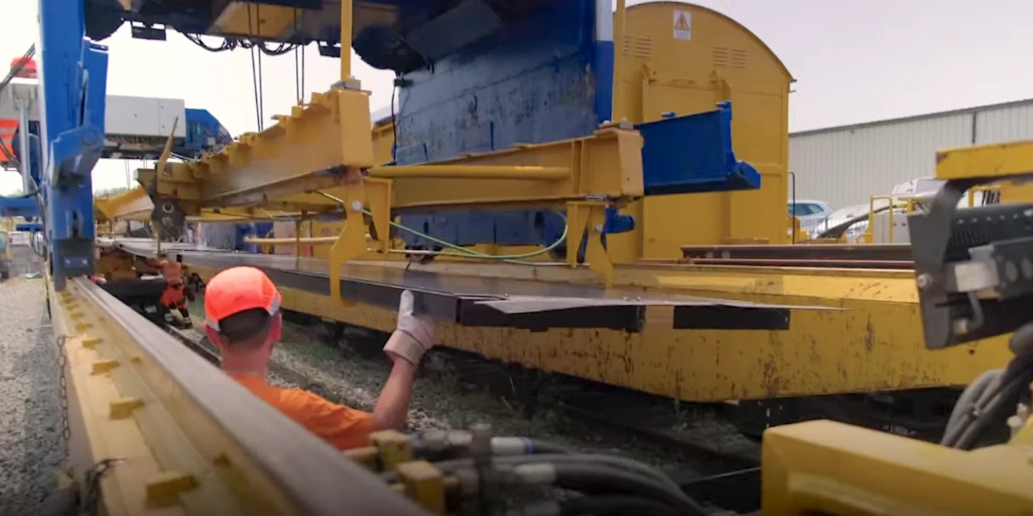 A construction worker in a red hard hat and orange shirt guides a large yellow industrial machine, installing solar panels on train tracks