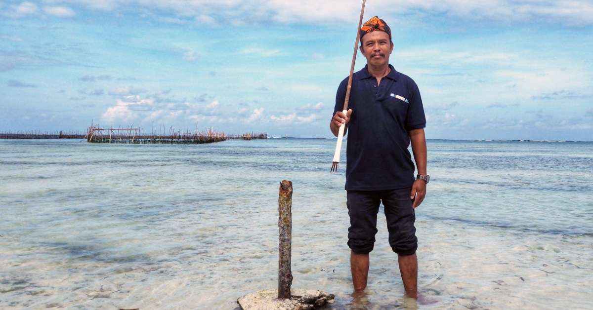 A man in a dark polo shirt and orange snorkel gear stands in shallow ocean water holding a wooden rake-like tool, with traditional fish traps visible in the background across the water.