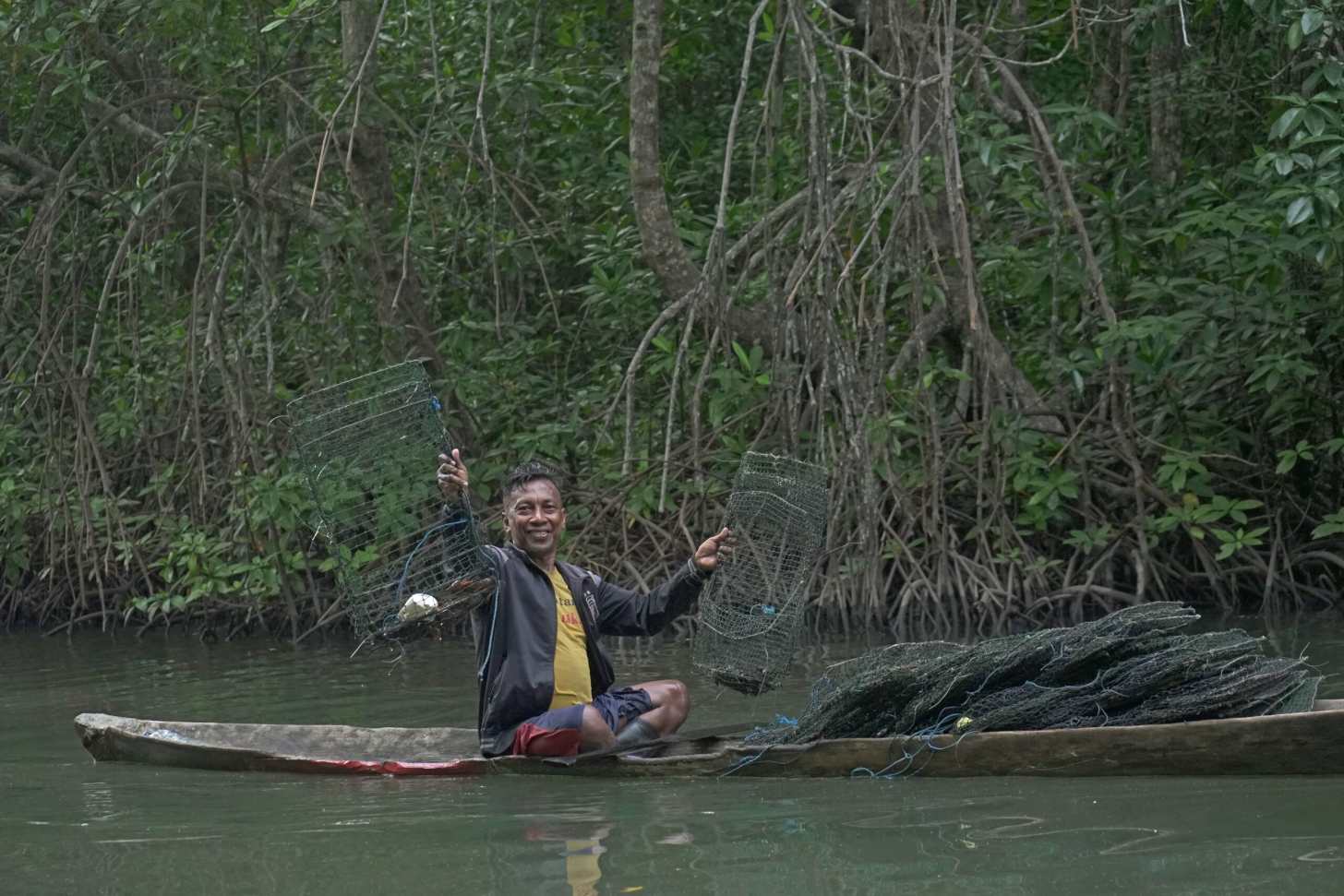 A fisherman smiles while holding up two fishing nets in shallow water surrounded by dense mangrove trees, displaying his catch from traditional fishing practices.