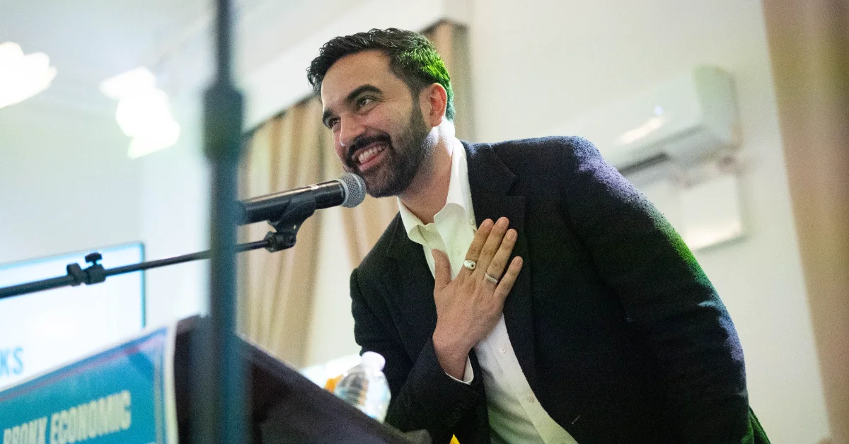 New York City Mayor Zohran Mamdani, wearing a dark blazer and white shirt, speaks into a microphone at a podium, smiling while gesturing with his hand to his chest.