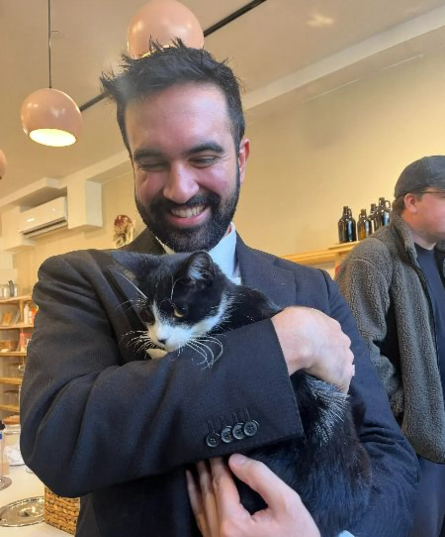 Zohran Mamdani wears a dark suit and tie and holds a black and white cat and smiles at it indoors, with another person visible in the background.