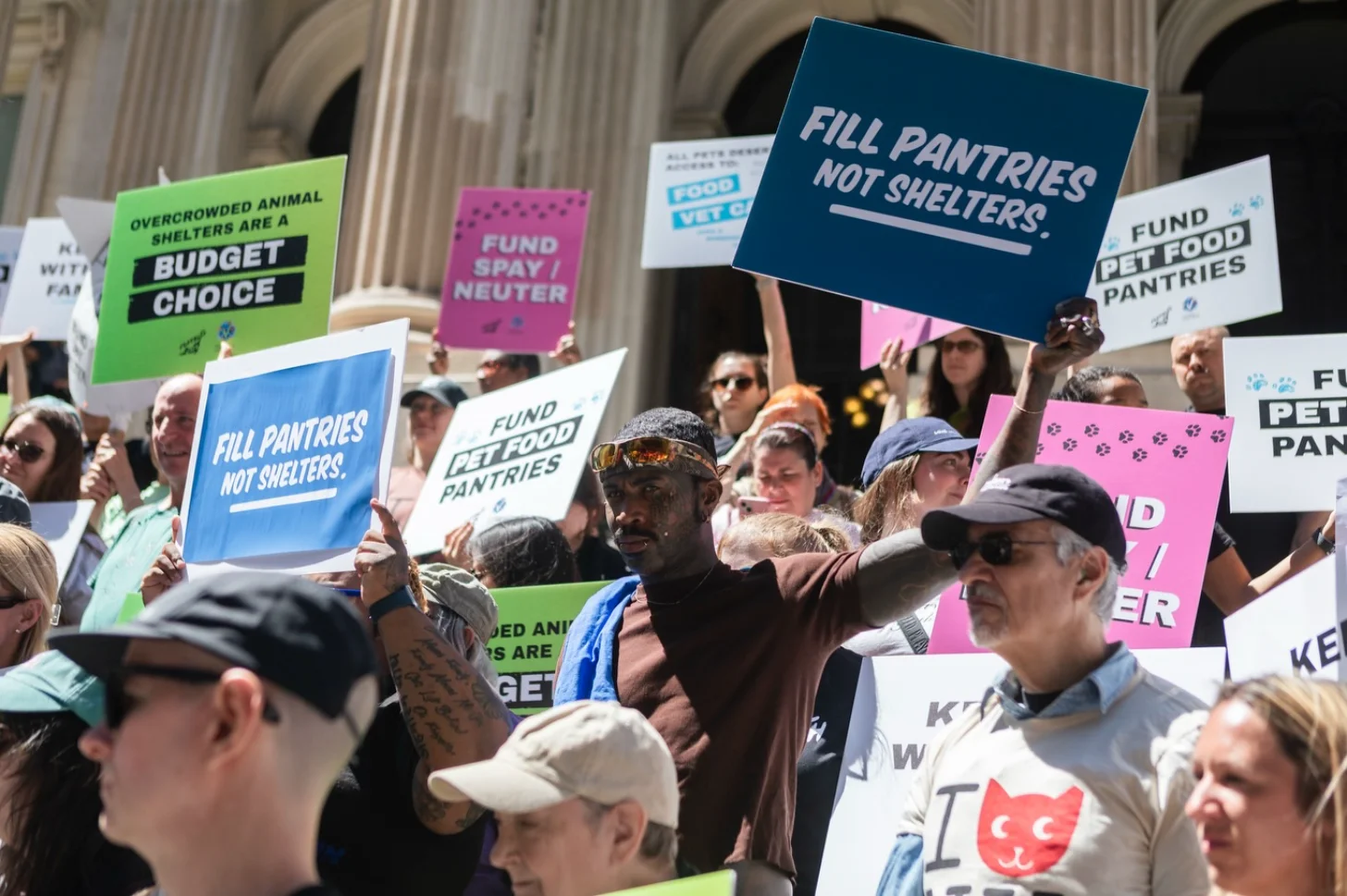 A crowd of people hold colorful signs outside a stone building. Signs read "FILL PANTRIES NOT SHELTERS," "FUND PET FOOD PANTRIES," "FUND SPAY/NEUTER," and "OVERCROWDED ANIMAL SHELTERS ARE A BUDGET CHOICE."