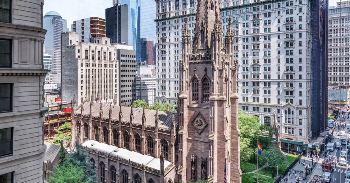 Gothic Revival church with spire and clock tower surrounded by modern skyscrapers, with construction scaffolding visible on its roof and a rainbow flag hanging on an adjacent building.