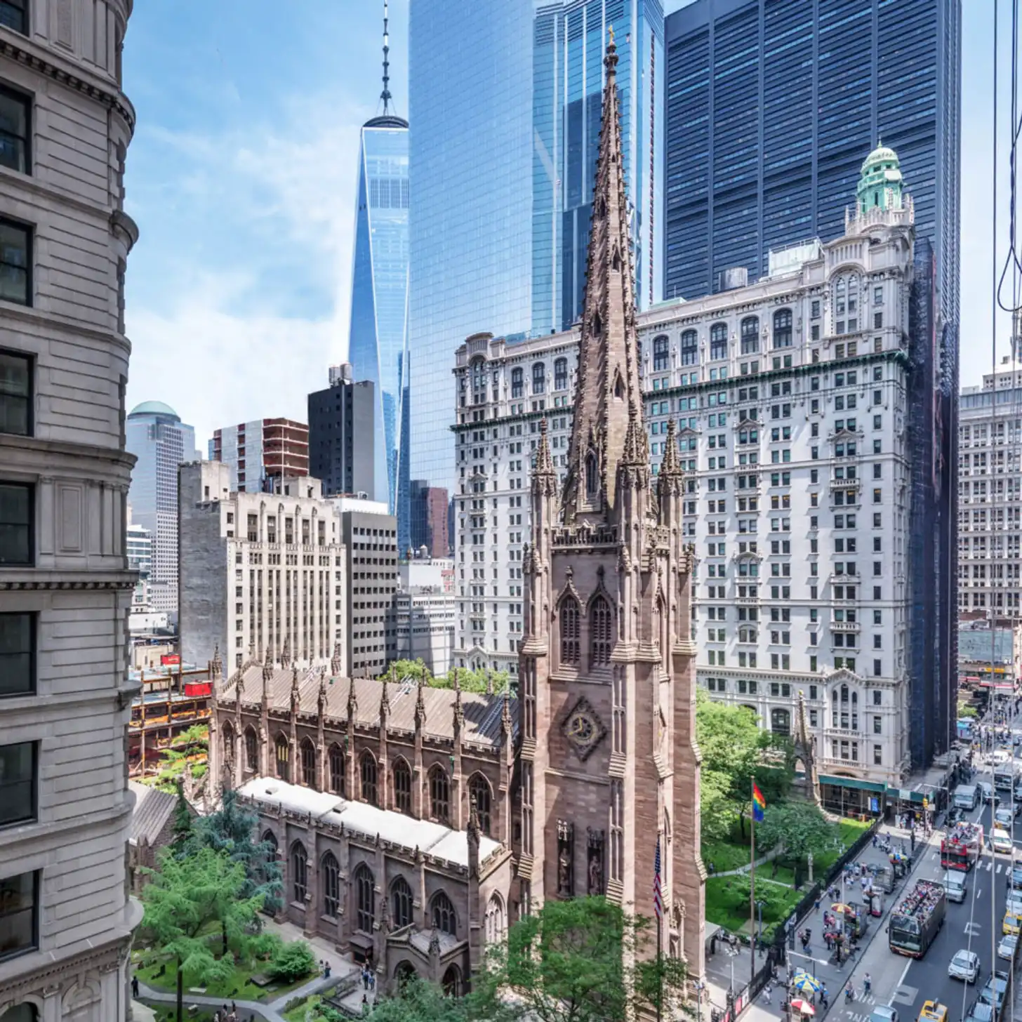 Gothic Revival church with ornate spire surrounded by modern glass skyscrapers, historic stone buildings, and street-level pedestrians and vehicles in an urban downtown setting.
