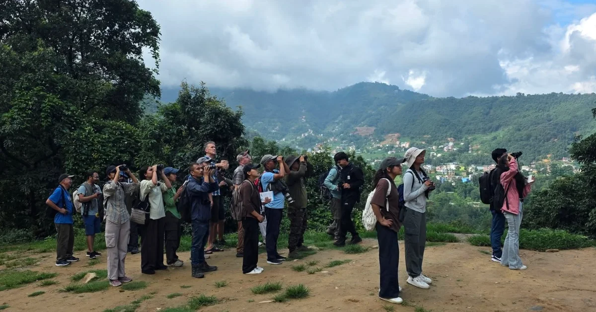 A group of people stand on a dirt overlook holding cameras and phones toward a mountainous valley with terraced settlements and lush green hillsides under a cloudy sky.