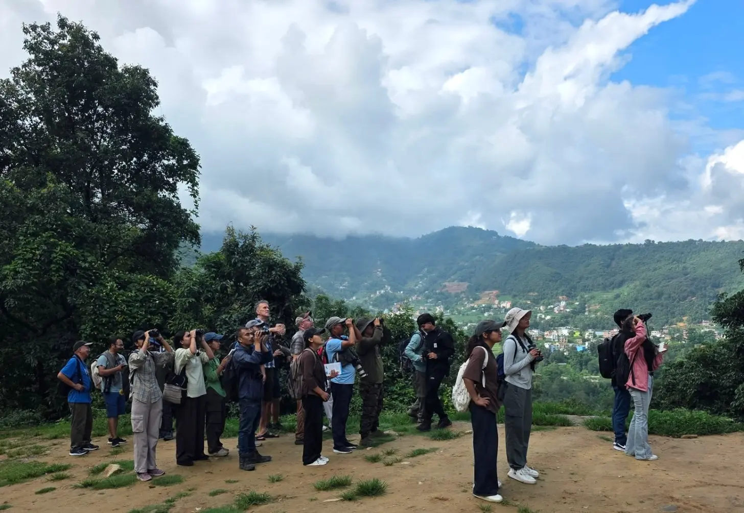 A group of people stand on a dirt overlook with mountains and a valley settlement visible in the background. Many hold cameras and binoculars aimed toward the landscape.