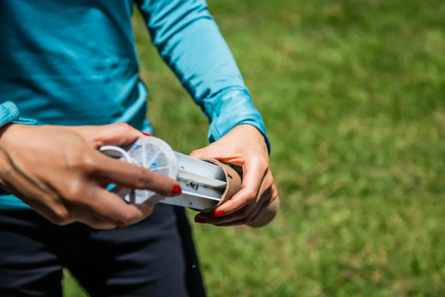 A person wearing a blue shirt holds a tube outdoors as they release mosquitoes. There is grass in the background, and the person has red nail polish visible on their fingernails.