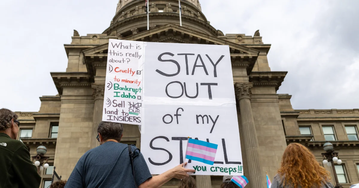 Protesters hold signs in front of a large stone government building. One sign reads "STAY OUT of my STALL you creeps!!!" with a transgender flag; another lists "Cruelty to minority," "Bankrupt Idaho," and "Sell land to our INSIDERS."