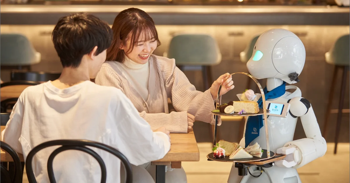 A white robot serves food on tiered trays to two people seated at a wooden table in a café. The woman on the left smiles while the robot, wearing a blue apron, presents the dishes.