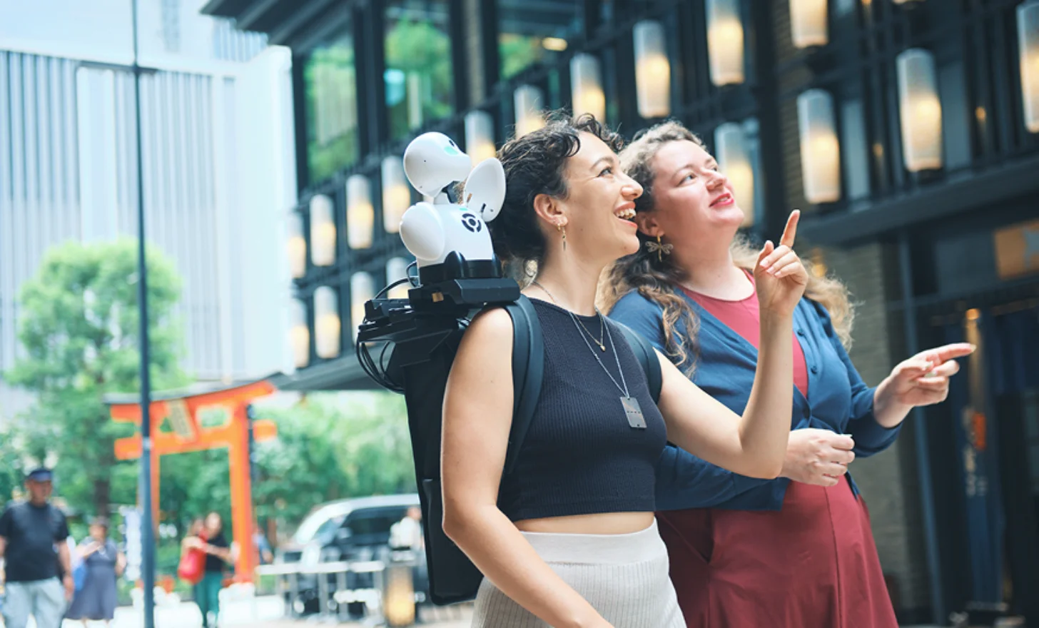 Two women stand outdoors looking upward and smiling, one wearing a black sleeveless top with a small robot on her shoulder, the other in a red and blue shirt. Behind them are modern buildings and urban street scenery.