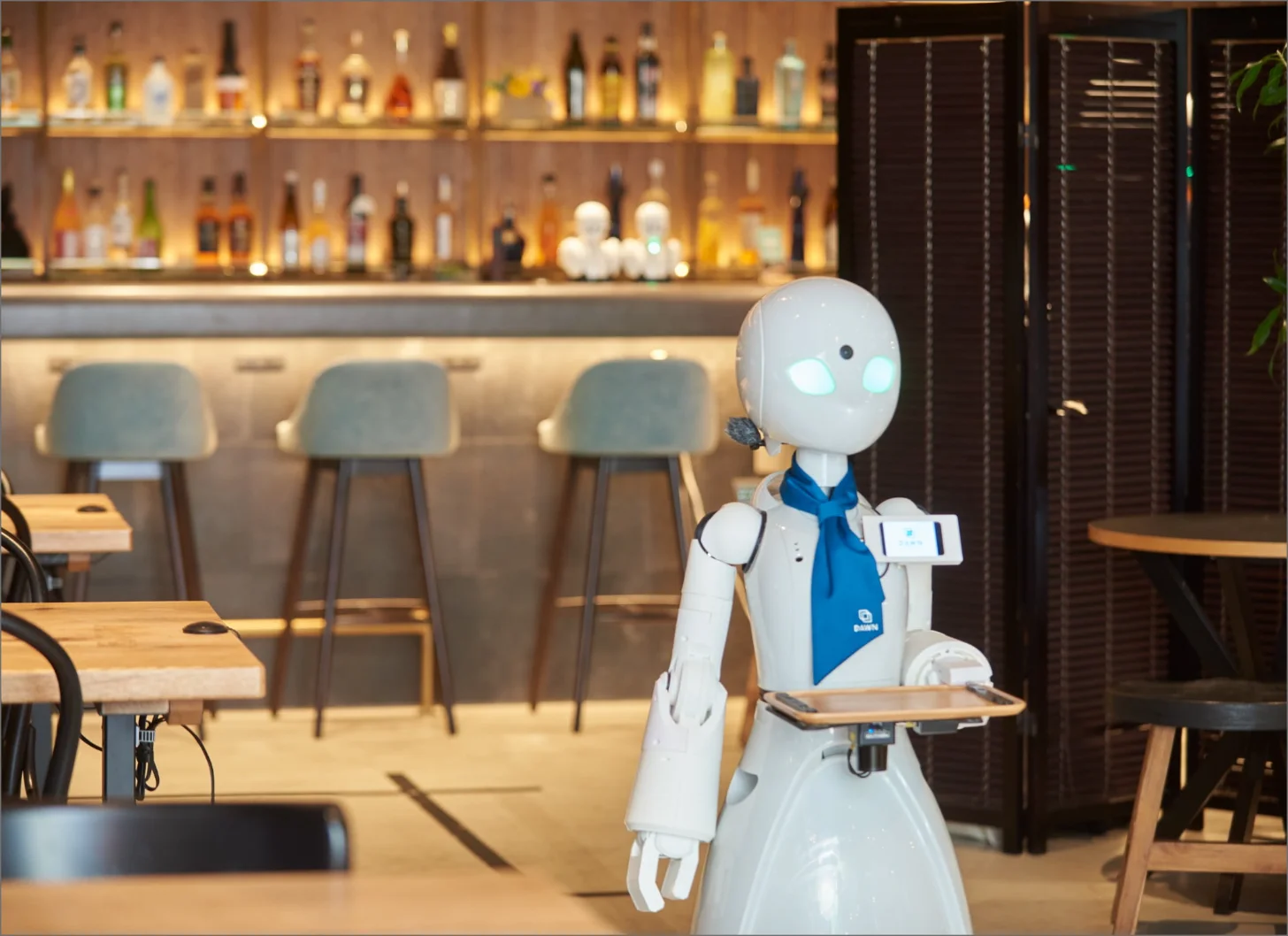 A white humanoid robot wearing a blue neckerchief holds a wooden serving tray in a bar or restaurant interior, with illuminated shelves of bottles visible behind the counter and empty tables in the foreground.