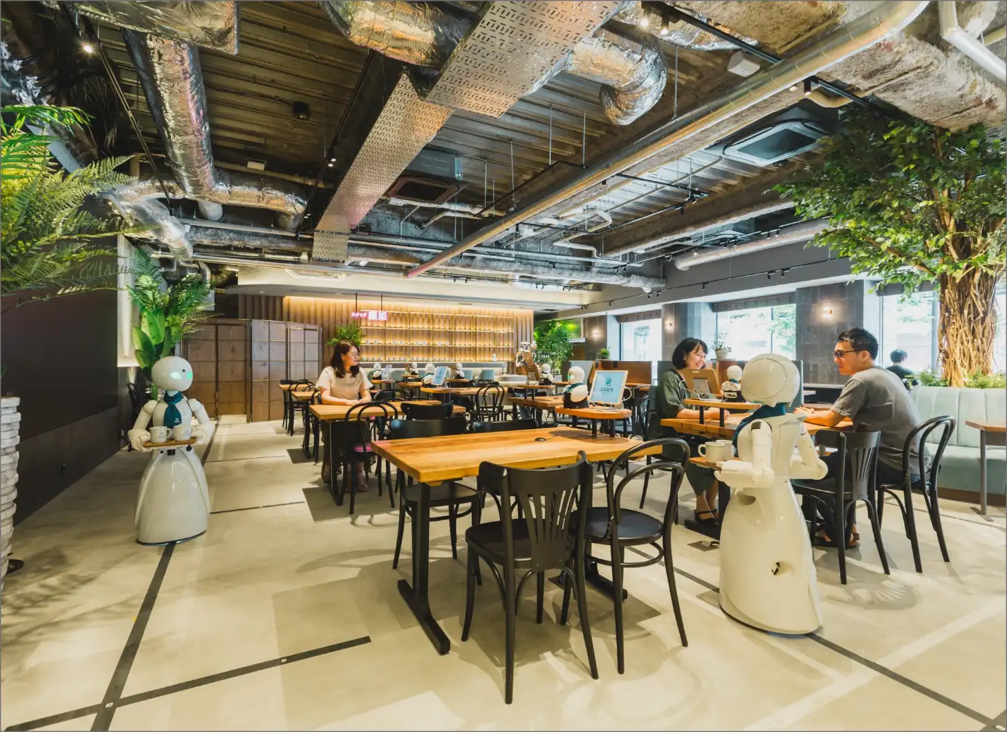 A modern restaurant interior with white dome-shaped robots stationed throughout, wooden tables with black chairs, and several diners seated at tables. Exposed ceiling with industrial elements, large windows, and living plant installations visible along the walls.