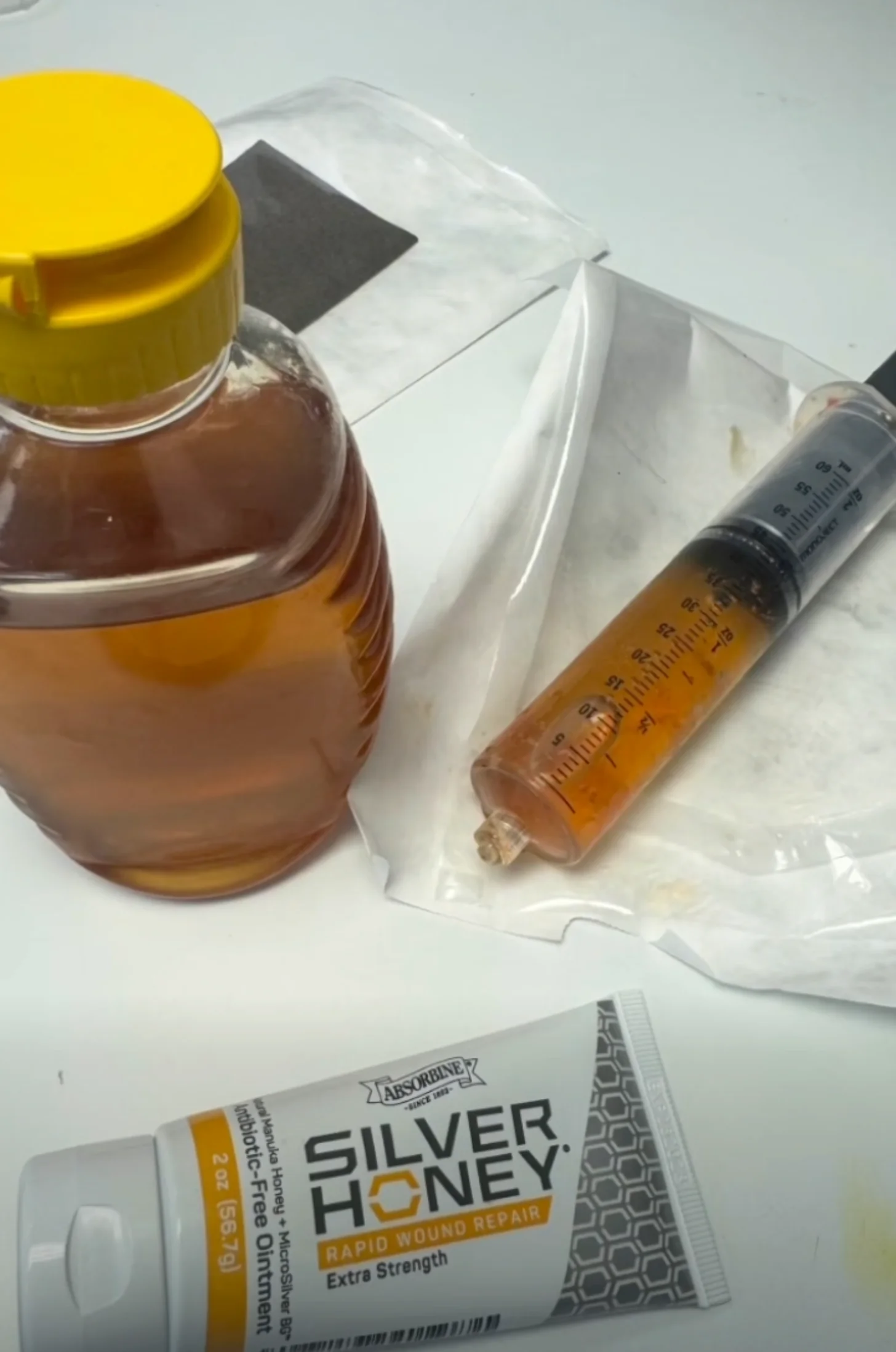 A bottle with a yellow cap containing amber liquid, a syringe filled with amber liquid, and a tube of Silver Honey Rapid Wound Repair Extra Strength arranged on white packaging and a light background.