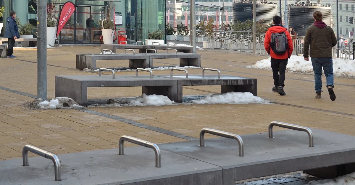 Two people in winter clothing run across a paved plaza with concrete seating and metal railings. Patches of snow are visible on the ground near modern furniture and storefronts with large windows.