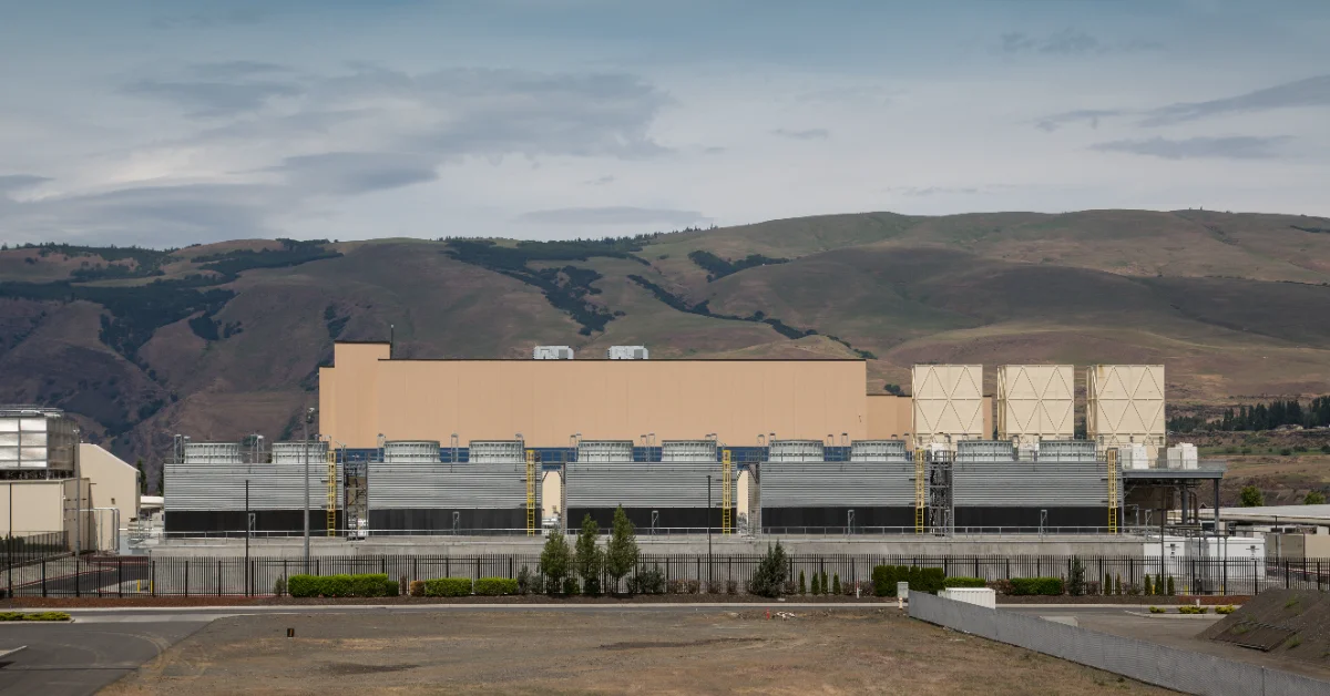 Large industrial facility with tan upper section and corrugated metal lower section, set against rolling hills with scattered forest in background.