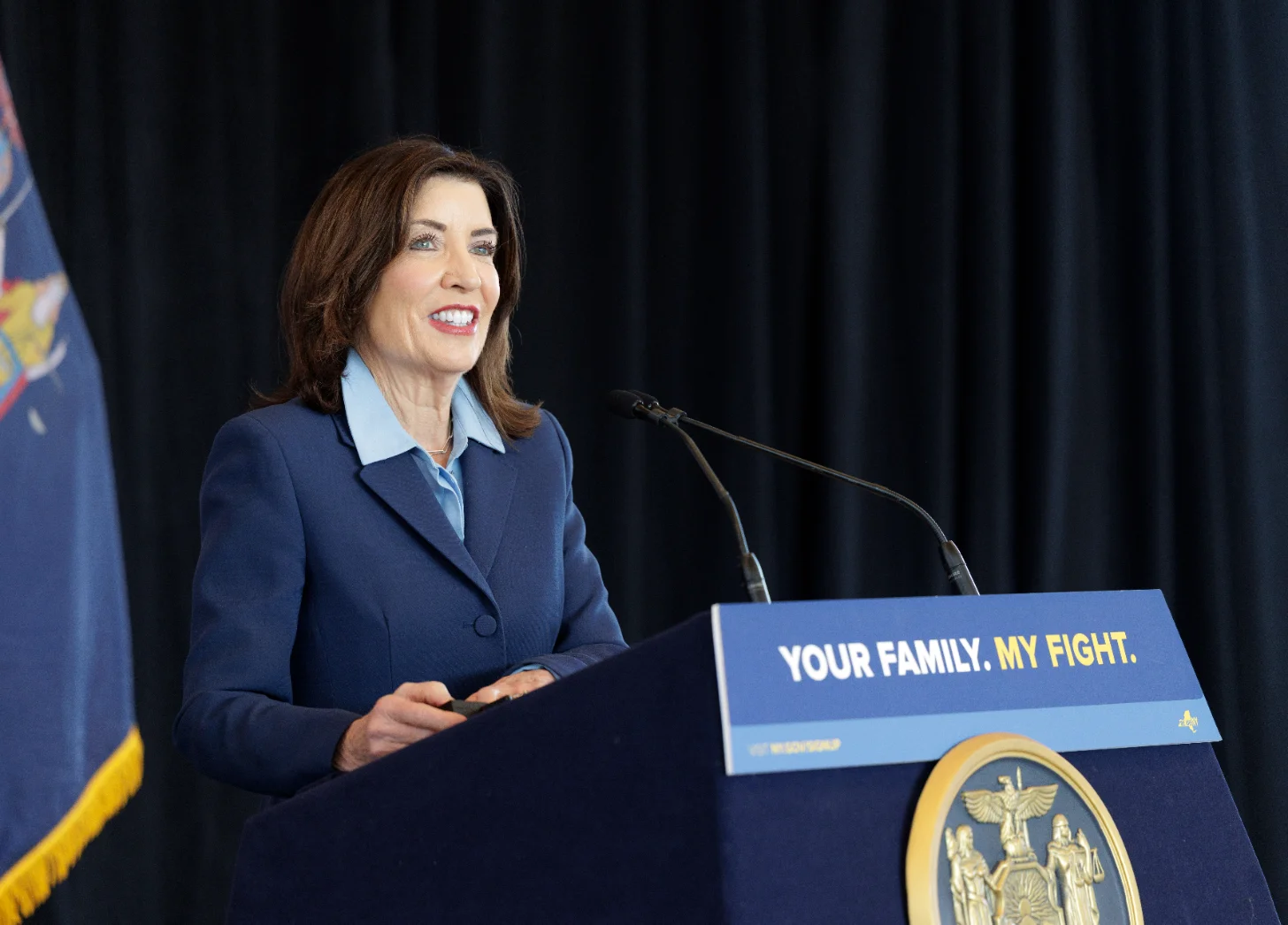 New York Governor Kathy Hochul in dark blue blazer speaking at podium with official seal. Podium sign reads "YOUR FAMILY. MY FIGHT." State flag visible in background.
