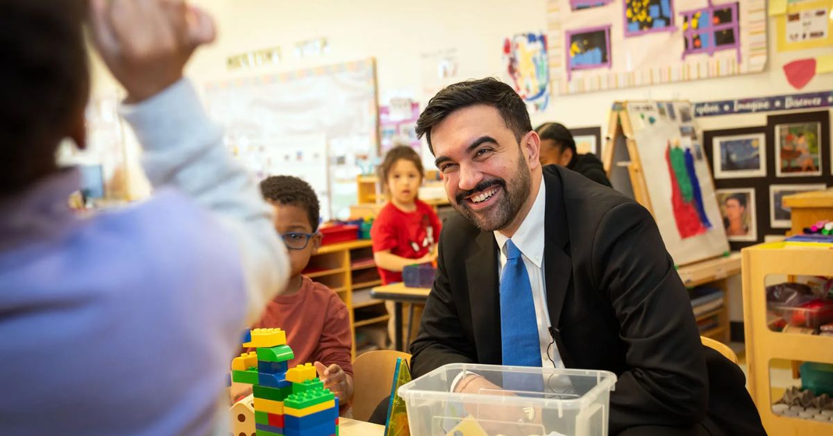 Mamdani sits in a classroom looking at a kid that's out-of-focus and out-of-frame