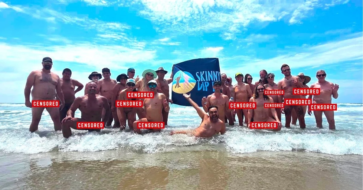 A large group of people standing in ocean waves at a beach with a blue sky above. A banner reading "SKINNY DIP" is visible among the group.