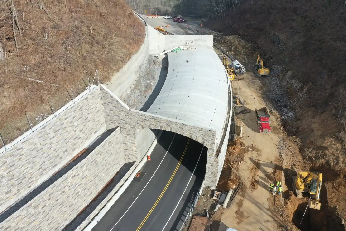 Aerial view of a tunnel entrance carved through a hillside with a stone facade, showing a two-lane paved roadway inside. Construction equipment and vehicles are visible on the road surface above and at an excavation site to the right.