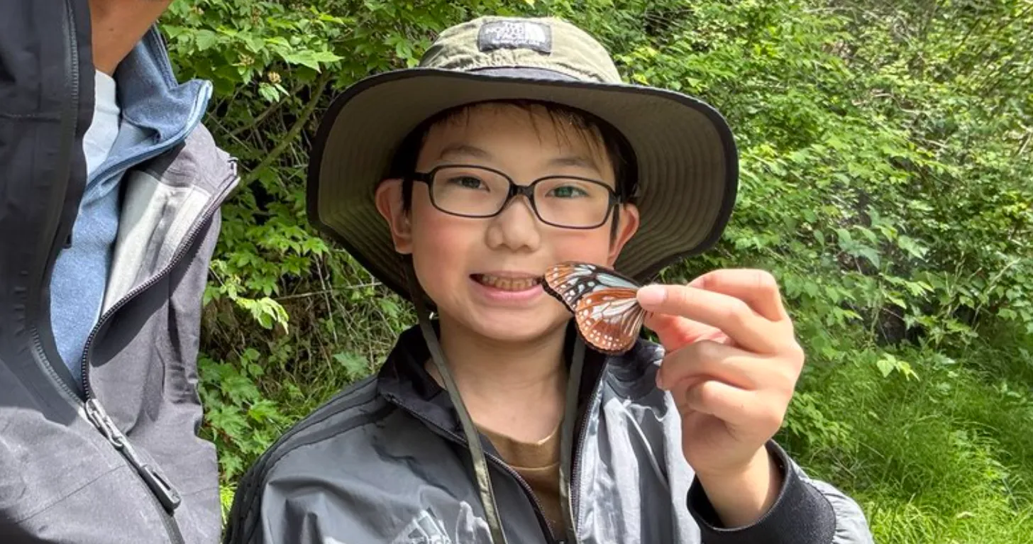 A young person wearing glasses and a wide-brimmed hat holds a monarch butterfly on their finger, smiling at the camera. Dense green foliage is visible in the background.