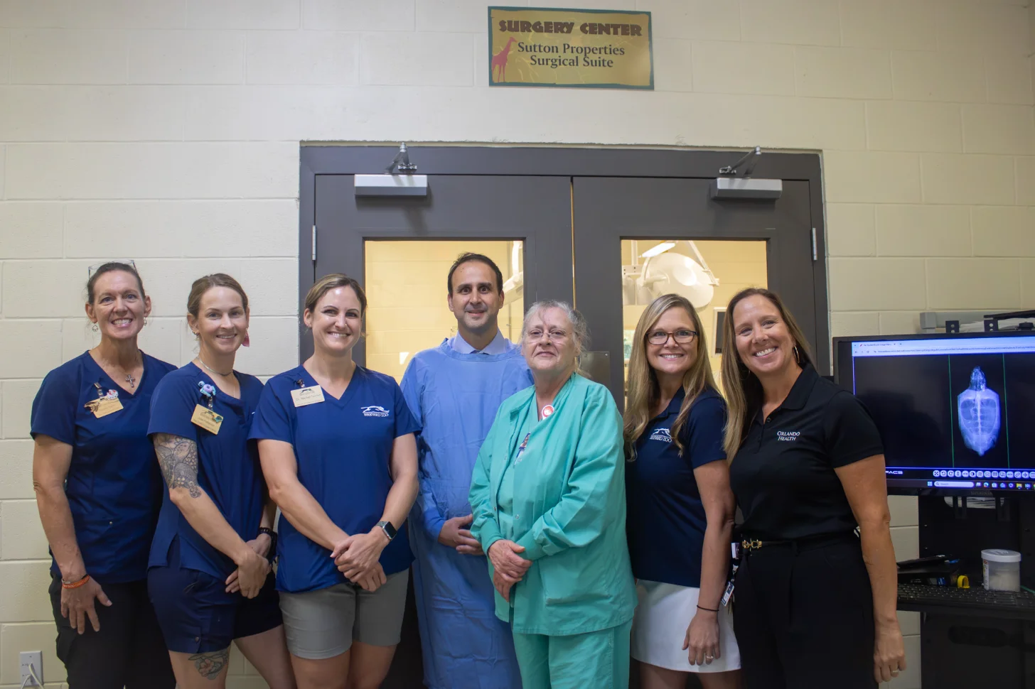 Seven medical professionals in scrubs stand together in front of a doorway labeled "Surgery Center, Sutton Properties Surgical Suite." A monitor displaying medical imaging is visible on the right.