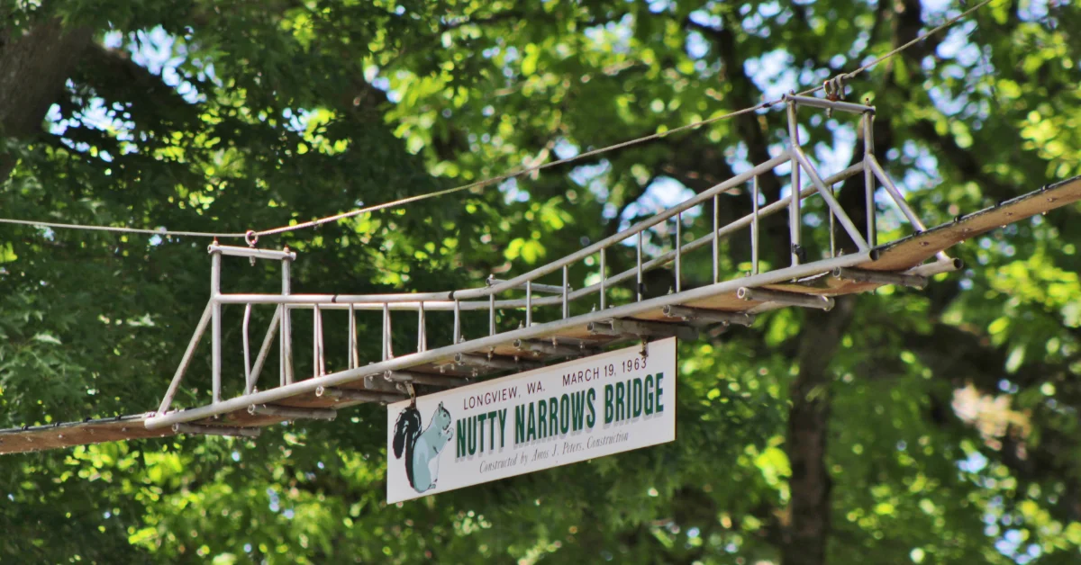 A suspension footbridge with metal railings and wooden planks spans between trees. A white sign reads "LONGVIEW, WA MARCH 19, 1963 NUTTY NARROWS BRIDGE" with an illustration of a squirrel.