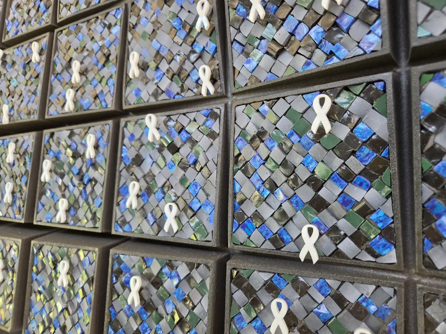 White awareness ribbons attached to framed mosaic tiles in shades of blue, green, gray, and black arranged in a grid pattern.