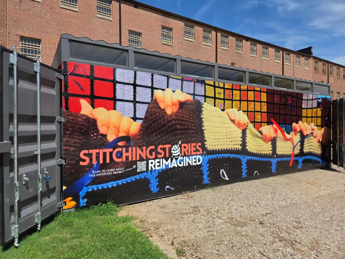 A shipping container with a mural displaying colorful knitted textile patterns and the text "STITCHING STORIES REIMAGINED" with a QR code, positioned against a brick building.