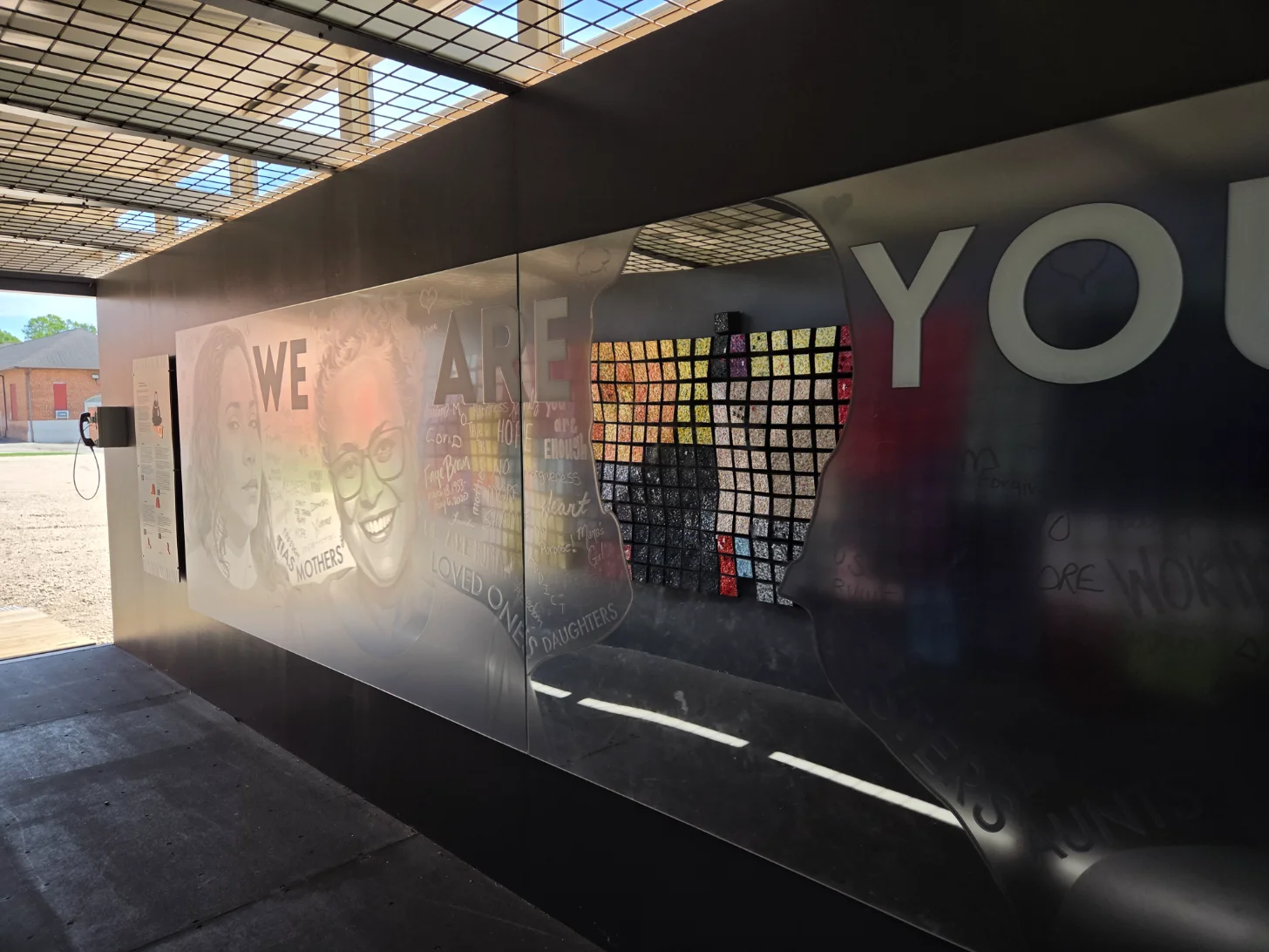 A dark architectural structure with glass panels displays text reading "WE ARE YOU" and features a grid of colorful photographs mounted on an interior wall. Sunlight streams through a gridded ceiling above.