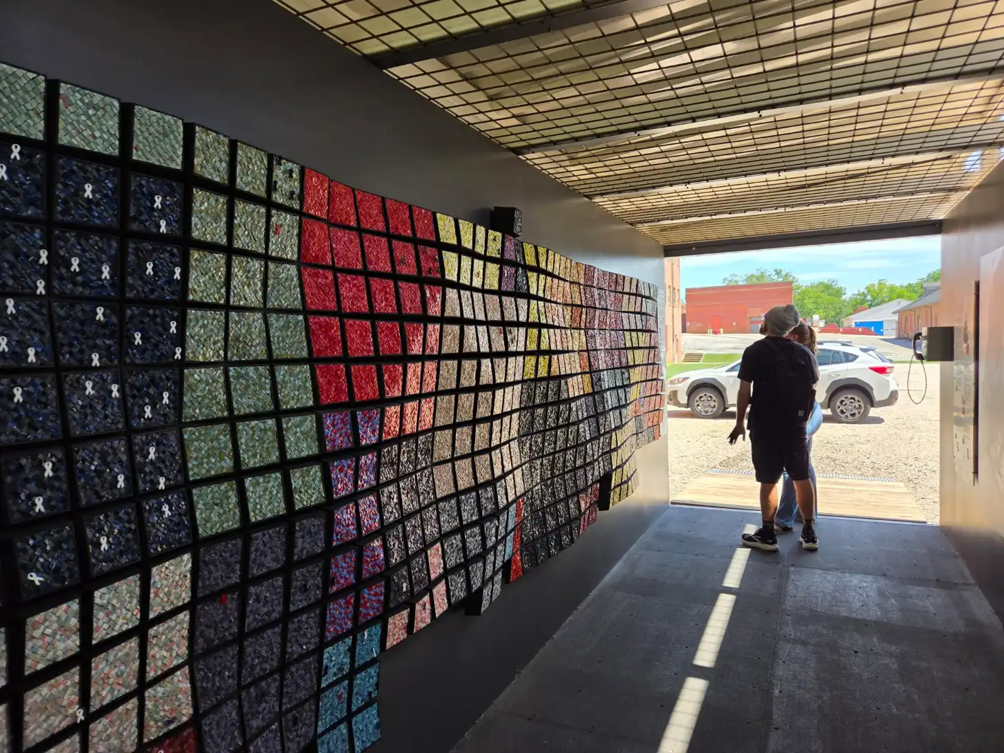 A person stands in a covered structure viewing a large wall display of fabric swatches arranged in a grid pattern, with colors ranging from dark blue and red to yellow and cream. A parking lot with vehicles and buildings are visible through the open entrance.