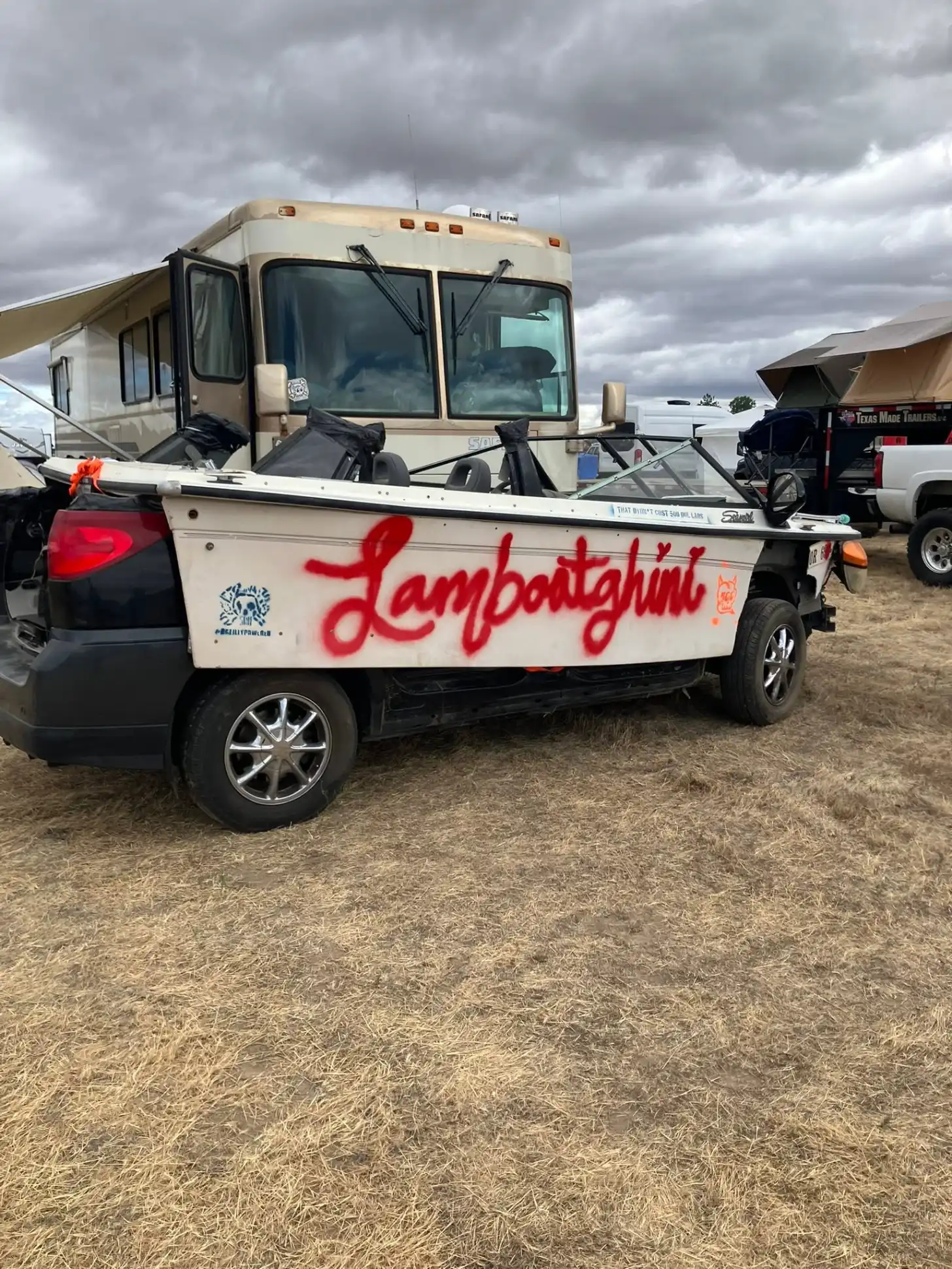 A pickup truck with "Lamborghini" written in red script on its white bed is parked at an RV campground under cloudy skies, with a tan motorhome visible behind it.
