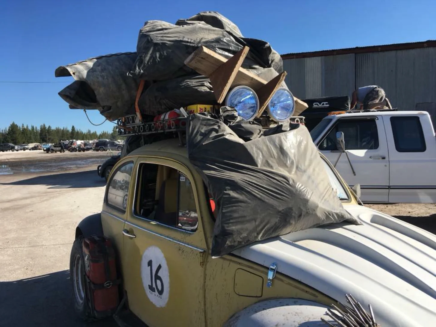 A yellow Volkswagen Beetle with the number 16 painted on its side carries an oversized fabric-wrapped figure with blue headlights and a brown box structure on its roof in a parking lot near a beach.