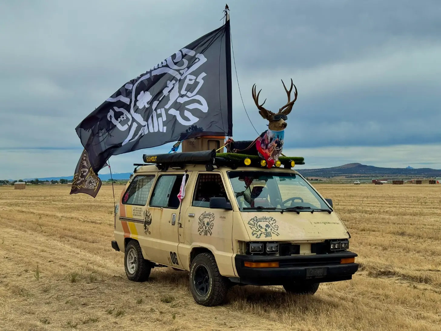 Tan minivan parked in an open field with a black flag flying from its roof rack and a large deer head sculpture mounted on top.