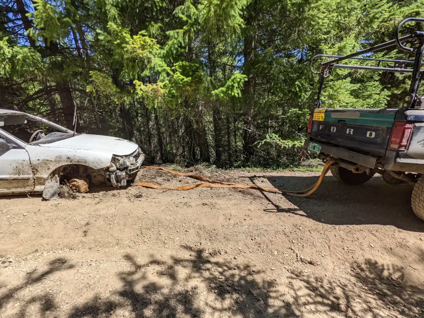 A white damaged sedan is being towed by a green Ford pickup truck on a dirt road lined with evergreen trees. An orange tow cable connects the two vehicles.