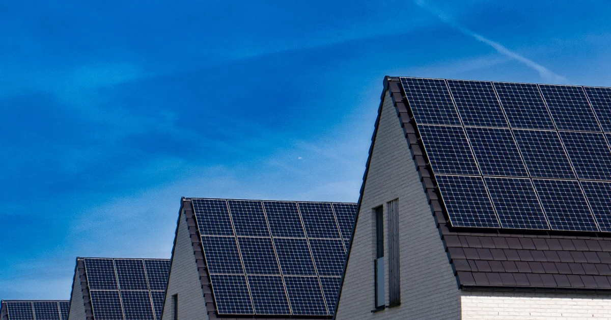 Row of residential houses with solar panel arrays mounted on their pitched roofs against a clear blue sky.