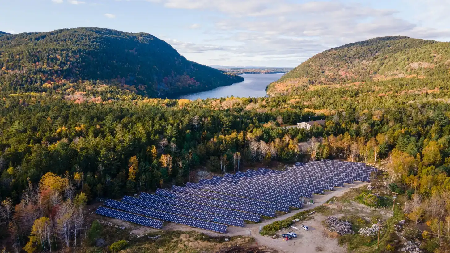 Large solar panel array in a forested valley with a lake visible between mountains in the background during autumn, with a house and vehicles at the facility.