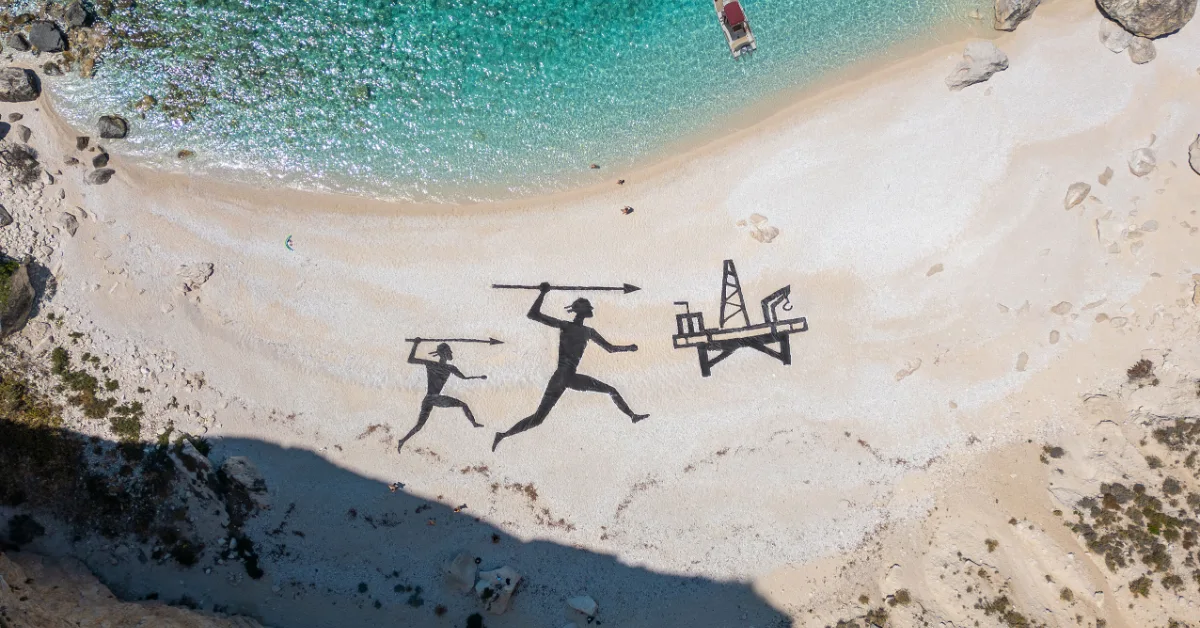 Aerial view of a beach with two silhouetted figures holding spears and an oil derrick painted in black on the sand, with turquoise water and a red boat visible above.