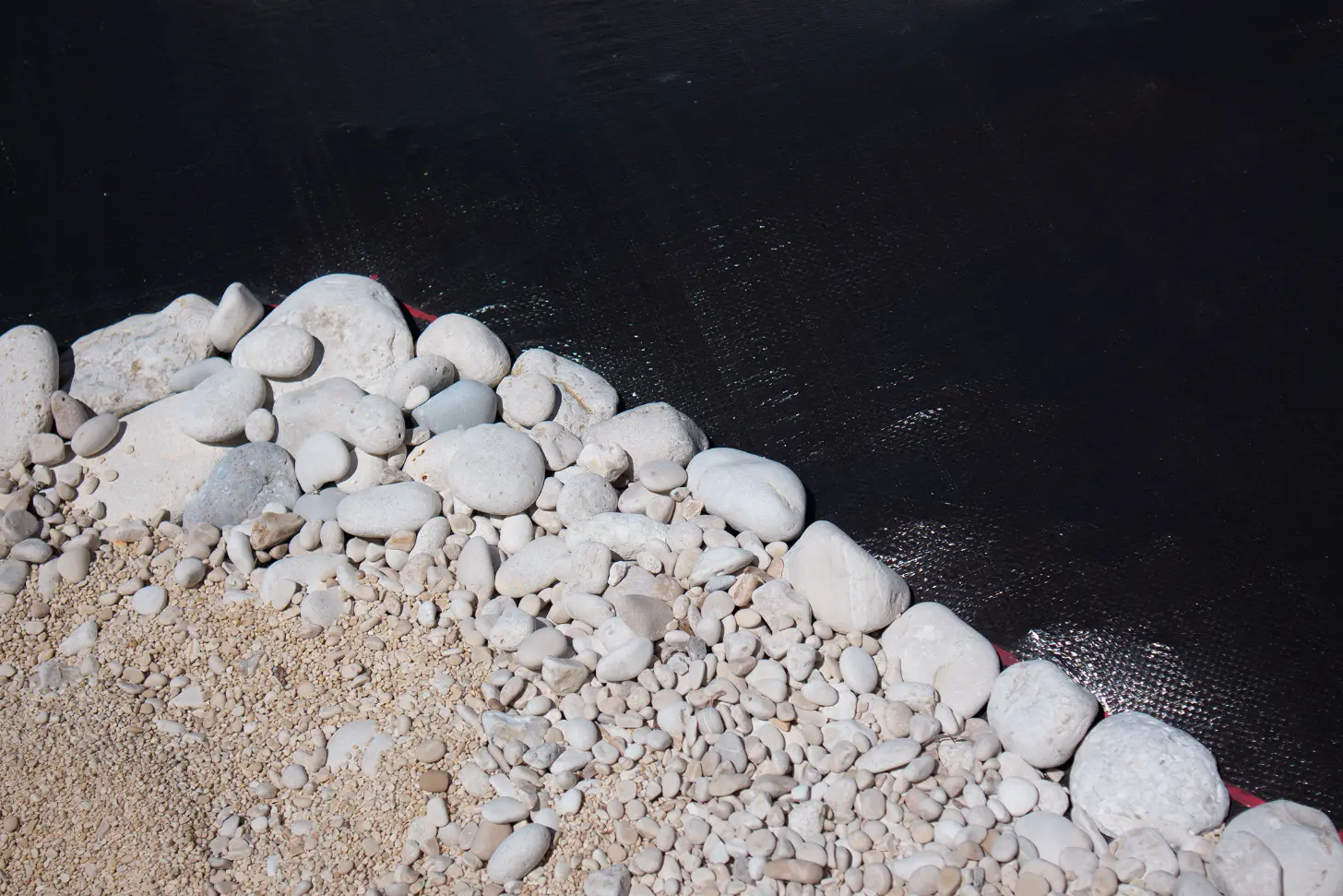White and tan rounded stones and pebbles line a dark fabric edge, with larger boulders visible along the shoreline.