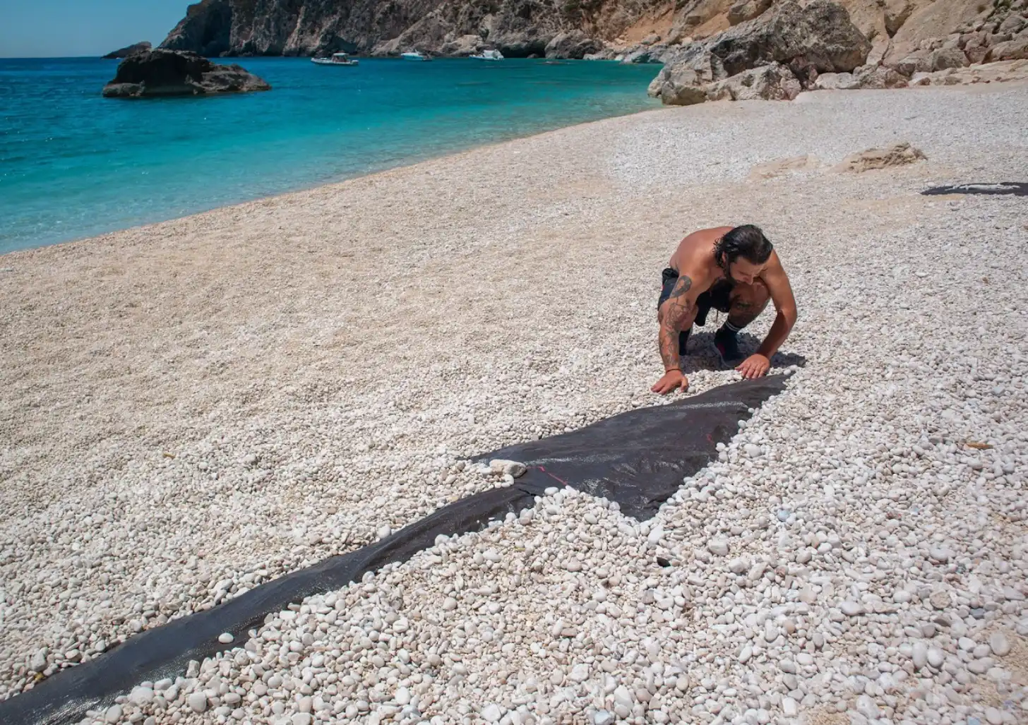 A man crouches beside a dark ray lying on a pebbly beach with turquoise water and rocky cliffs visible in the background.